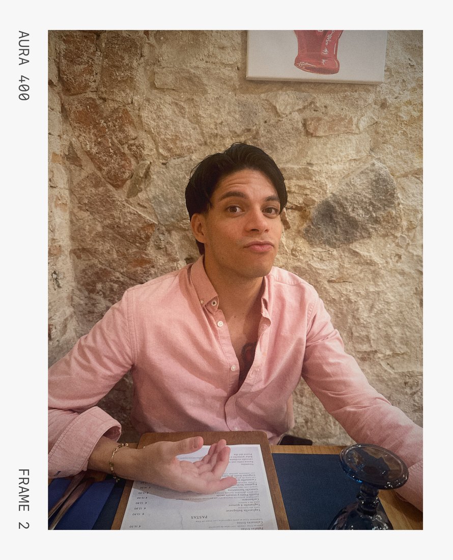 A man in a pink shirt reading a restaurant menu against a stone wall, framed with SloSta's AURA 400 film stock